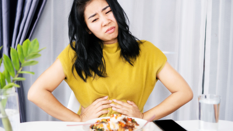 Junge Frau sitzt vor einem Teller voller Essen, hat Bauchschmerzen.