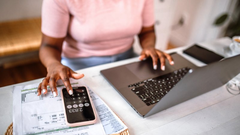 Frau sitzt am Schreibtisch mit ihrem Laptop und Handy und plant Haushaltsfinanzen.