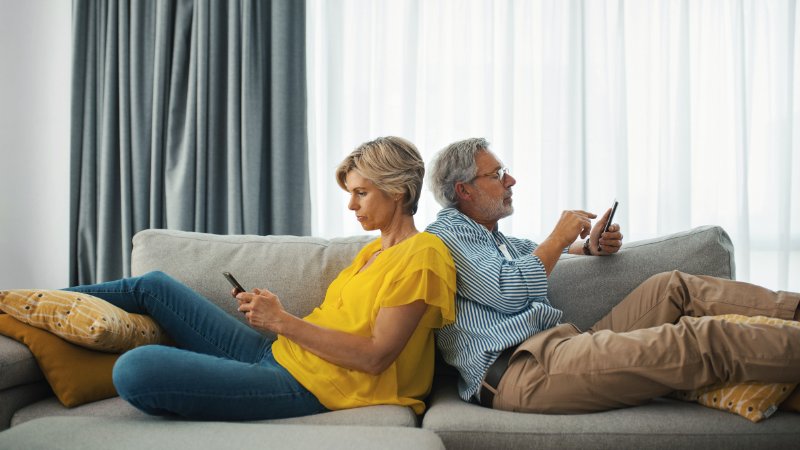 Ein älteres Ehepaar sitzt Rücken an Rücken auf dem Sofa. Beide schauen auf ihr Handy
