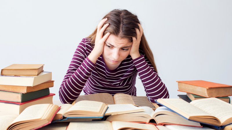 Junge Studentin sitzt verzweifelt beim Lernen vor vielen Büchern.